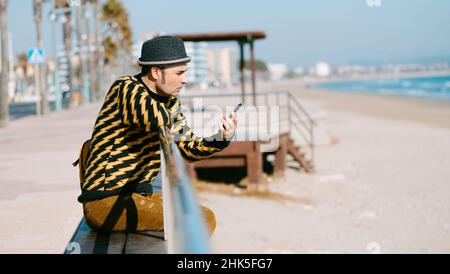 un uomo di mezza età, che indossa abiti casual moderni e colorati, guarda il suo smartphone, seduto all'aperto accanto alla sabbia di una spiaggia urbana Foto Stock