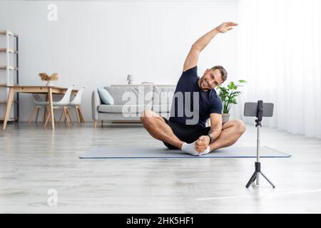 Allegro ragazzo caucasico muscoloso millenario in uniforme seduto sul pavimento su tappeto in posizione lotus, praticando yoga Foto Stock