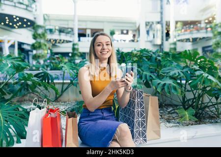 La giovane donna consumatore nel centro commerciale naviga chiacchierando e utilizza uno smartphone. La donna in piedi con un telefono cellulare nelle sue mani nel centro commerciale. Al coperto Foto Stock