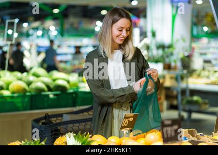 La giovane donna sceglie e sceglie in sacchi ecologici di verdure o frutta arance nel supermercato. Cliente femminile in piedi un negozio di alimentari vicino al banco bu Foto Stock