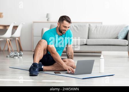 Sorridente allegro millenario muscoloso caucasico maschio blogger seduto su tappeto con bottiglia d'acqua e computer Foto Stock