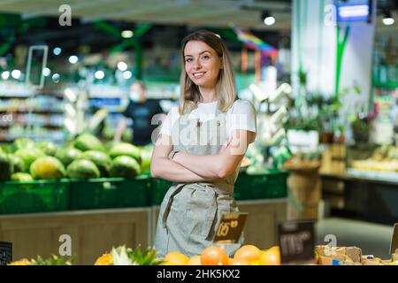 Ritratto giovane donna lavoratore in una sezione vegetariana supermercato in piedi sorridendo con le braccia incrociate. Amichevole simpatica femmina che guarda la fotocamera in una fru Foto Stock