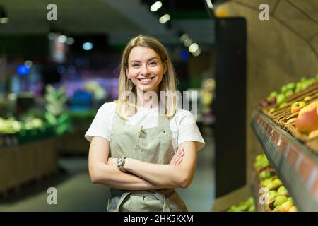 Ritratto giovane donna lavoratore venditore in una sezione di verdure supermercato in piedi in braccia incrociate. Greengroer femmina guardando la macchina fotografica in negozio di frutta mar Foto Stock