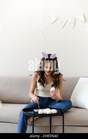 Bella giovane donna in orecchie di coniglio dipinge uova per pasqua. Foto Stock