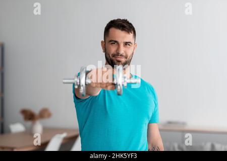 Allegro giovane muscoloso europeo uomo fare braccio esercizi, sollevamenti dumbbell guardare la macchina fotografica in soggiorno Foto Stock