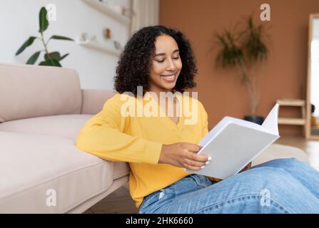 Giovane donna nera sorridente che legge il notebook, seduto sul pavimento, preparandosi per l'esame o la riunione d'affari a casa Foto Stock