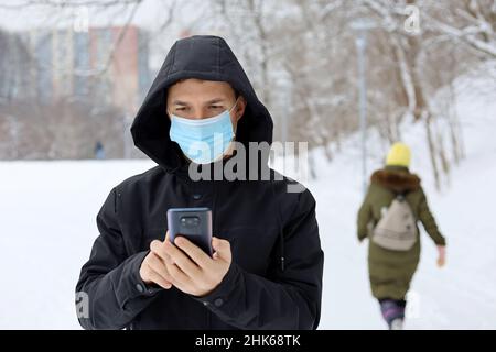 Protezione da coronavirus, uomo in maschera medica su una strada invernale della città con smartphone in mano. Concetto di sicurezza durante il covid-19 pandemia, tempo di neve Foto Stock