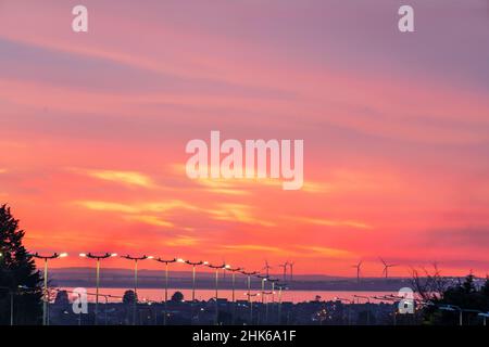 Cielo notturno nuvoloso rosso e giallo vivido dopo il tramonto sulla lontana isola Kent di Sheppey vista da Herne Bay. Alcune grandi turbine eoliche all'orizzonte e una fila di luci di strada del A299 Thanet Way che conduce al paesaggio. Foto Stock