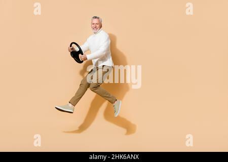 Vista a tutta lunghezza del corpo di un attraente uomo dai capelli grigi e allegri che salta tenendo il volante isolato su sfondo color pastello beige Foto Stock