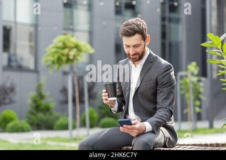 Bell'uomo d'affari felice bearded seduto su panca con caffè utilizzare il telefono cellulare navigare smartphone in mani legge buone notizie e gioioso sorriso. Outs Foto Stock