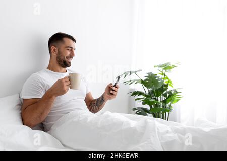Sorridente uomo europeo muscoloso millennial bere caffè, chiacchierare sullo smartphone e godersi la buona mattina Foto Stock