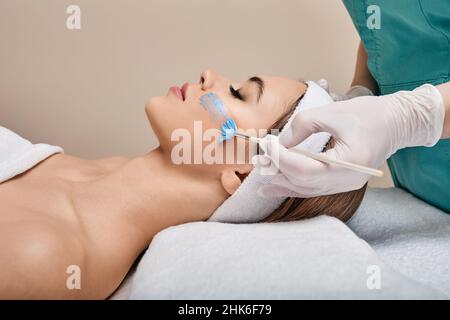 L'estetista applica la maschera di cura al viso della donna utilizzando la spazzola cosmetologica. cura della pelle del viso con maschera cosmetica presso la clinica di bellezza Foto Stock