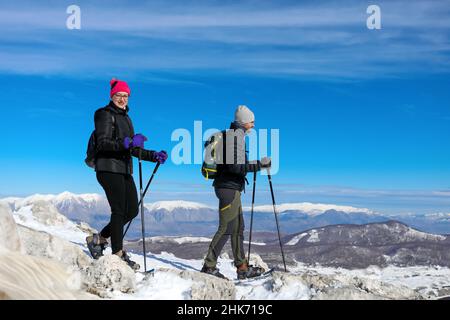 Due escursionisti camminano con le racchette da neve fino alla vetta innevata, circondata da un paesaggio incontaminato e da catene montuose innevate. In una bella giornata invernale. Foto Stock