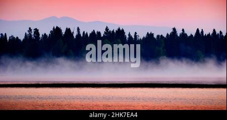 All'alba o al tramonto su un lago con nebbia che salgono dal acqua e alberi di pino montagne in strati di sfondo Foto Stock