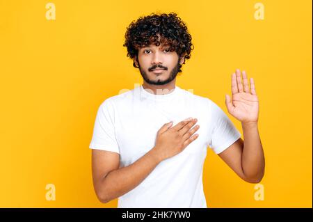 Onesto orgoglioso curly-paired giovane adulto indiano o arabo uomo in bianco casual t-shirt, tenendo la mano sul petto e alzando la palma, dando promessa, giuramento, in piedi su isolato sfondo arancione Foto Stock