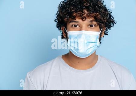 Prevenzione delle infezioni. Primo piano di un ragazzo curly indiano o arabo giovane adulto, con una maschera medica protettiva sul suo volto, guardando la fotocamera mentre si trova su un isolato sfondo blu Foto Stock