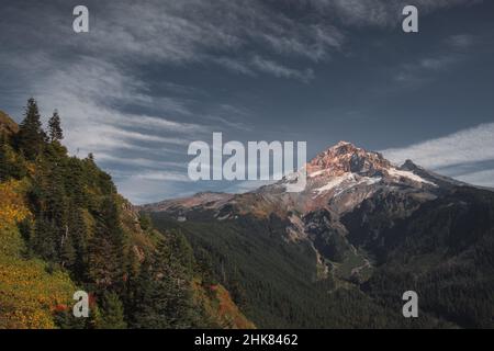 Una nube lenticolare abbraccia la cima del monte Hoot coperto di neve in uno splendido tramonto in autunno. Con un cielo perlopiù blu c'è una serie di vette di montagna Foto Stock