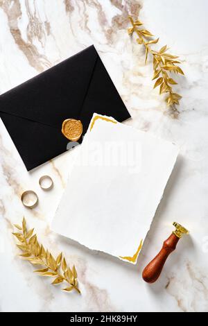 Mockup di cancelleria di nozze. Carta di invito per matrimoni vuota con busta nera, sigillo in cera, anelli dorati, foglie su sfondo oro marmo. Disposizione piatta Foto Stock