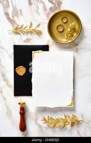 Set di articoli da cancelleria per matrimoni di lusso. Mockup carta di invito in bianco con busta nera, sigillo in cera, anelli dorati, foglie floreali su tavolo da scrivania in marmo oro Foto Stock