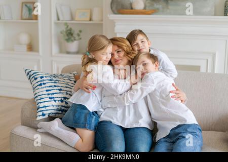 I bambini carini abbracciano, la mamma eccitata che mostra amore e affetto, la madre sorridente e la figlia e due figli in età scolare che si divertono a casa, abbracciando la condivisione intimo momento tenero insieme Foto Stock