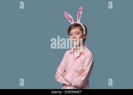 Un giovane ragazzo in camicia rosa con orecchie di coniglietto pasquale sulla testa si pone allegro e gesticula con le mani in studio su uno sfondo blu. Felicità e gioia dalle vacanze primaverili Foto Stock