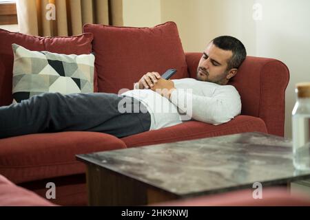 Foto ravvicinata di un uomo che soffre di narcolessia che dorme sul divano mentre tiene in mano lo smartphone. Foto Stock