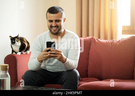Bell'uomo dai capelli corti che tiene lo smartphone e si siede sul divano con il gatto. Spazio di copia Foto Stock