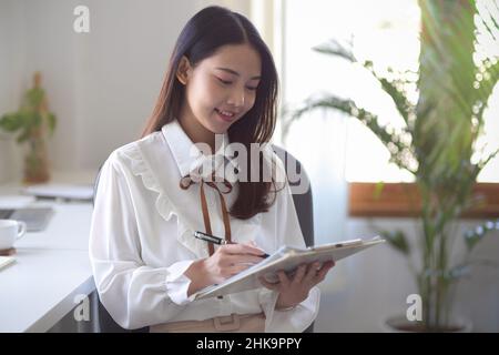 Piuttosto giovane donna asiatica ufficiale o imprenditore di affari che firma la sua firma sul documento nella sua stanza d'ufficio. Foto Stock