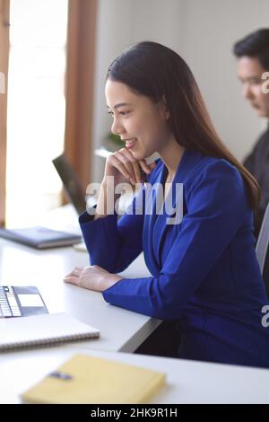 Vista laterale, Ritratto di una giovane donna d'affari asiatica di successo in abito elegante seduto alla sua scrivania, lavorando sul laptop e pensando a una nuova idea di business. Foto Stock
