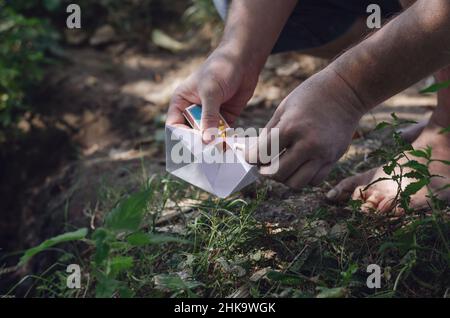 Un uomo adulto a piedi nudi illumina una barca di carta. Mani che tengono una barca bianca per bambini origami e un fiammifero ardente. Ora legale. Trick selettivo. All'esterno del Foto Stock