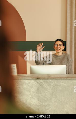 Un'allegra receptionist femminile dà il benvenuto ad un cliente alla reception. Felice amministratore femmina che agita la mano ad un cliente entrante mentre in piedi beh Foto Stock