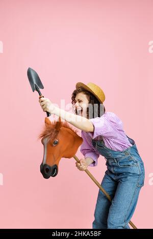 Ritratto comico di ragazza giovane allegra, giardiniere in tute denim e cappello che si divertono isolato su sfondo rosa. Concetto di lavoro, emozioni, agronomia Foto Stock