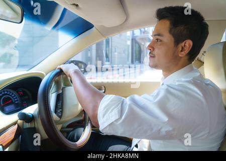 Il giovane uomo asiatico sorride e guarda la strada urbana mentre guida un'auto in città Foto Stock