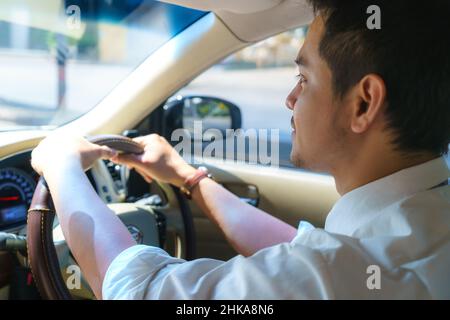 Il giovane uomo asiatico sorride e guarda la strada urbana mentre guida un'auto in città Foto Stock