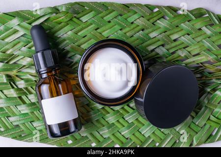 Olio viso in bottiglia contagocce nera e crema idratante aperta in vaso di vetro su foglia tropicale e sfondo di marmo. Concetto di cosmetici naturali. Branding Foto Stock