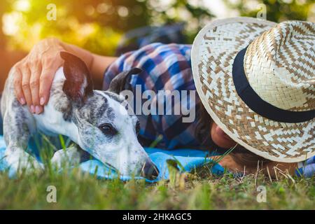 PET donna proprietario rilassarsi con il suo cane da frusta in giardino. Animale domestico carino che posa nell'erba. Donna che indossa un cappello di paglia. Foto Stock