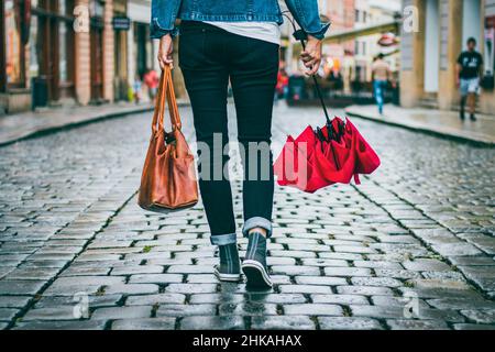 Donna che tiene la borsa di pelle e ombrello rosso e sta camminando sulla strada dopo la pioggia. Concetto di moda in città. Donna alla moda che indossa abiti casual. Foto Stock