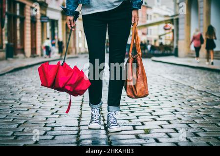 Donna che tiene la borsa di pelle e ombrello rosso e sta camminando sulla strada dopo la pioggia. Concetto di moda in città. Donna alla moda che indossa abiti casual. Foto Stock