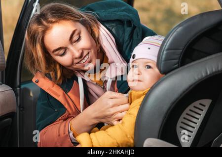 Sorridente giovane madre caucasica prepara il suo bambino carino per un viaggio in auto. Una donna allaccia le cinture di sicurezza in un seggiolino per bambini. Vista all'interno di un'auto. Foto Stock