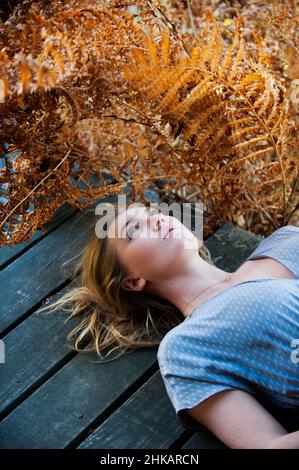 Giovane donna adagiata su portico di legno, circondata da felci, rivolta verso l'alto Foto Stock