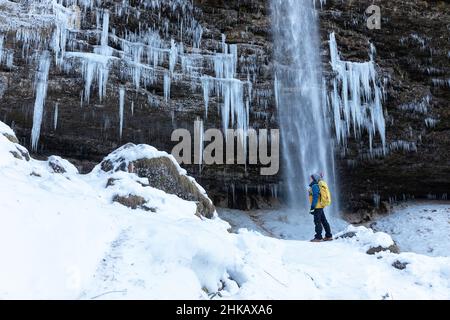Ragazzo con uno zaino in piedi di fronte alla cascata ghiacciata della cascata di Pericnik in inverno, Parco Nazionale del Triglav, Slovenia Foto Stock