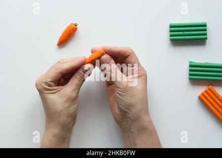 Le mani della donna che fanno l'argilla piccola del polimero della carota sullo sfondo bianco Foto Stock