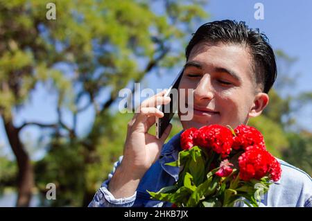 giovane latino caucasico ispanico innamorato parlando al telefono con il suo amante con gli occhi chiusi tenendo un bouquet di fiori rossi vicino al suo viso outdo Foto Stock