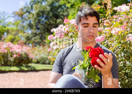 uomo caucasico in un giardino seduto sul pavimento che contempla un bel bouquet di fiori rossi il giorno di san valentino, uomo innamorato, immagine con spazio copia Foto Stock