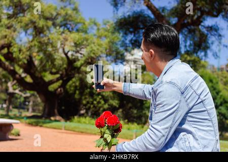 giovane ispanico latino argentino caucasico seduto in un parco con alberi e vegetazione, facendo videochiamata con il suo telefono, comunicando con il suo g Foto Stock