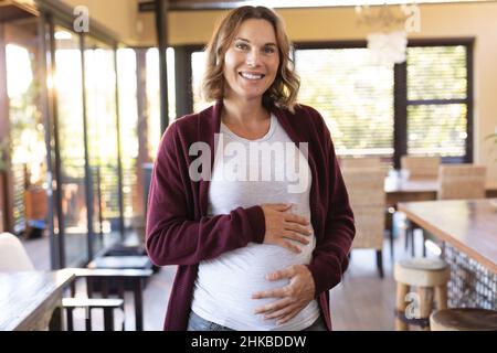 Ritratto di donna incinta caucasica che tocca il ventre e sorride a casa Foto Stock