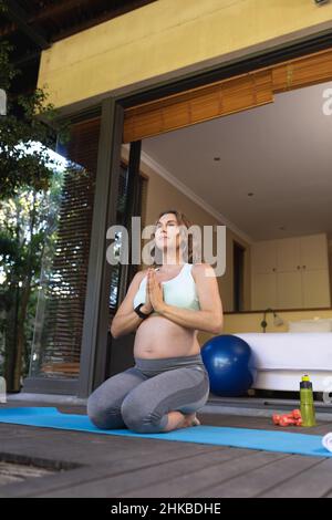 Donna incinta caucasica che medita ed esegue yoga seduto sul tappeto yoga a casa Foto Stock