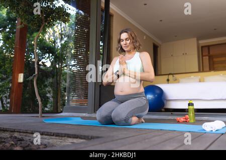 Donna incinta caucasica che medita ed esegue yoga seduto sul tappeto yoga a casa Foto Stock