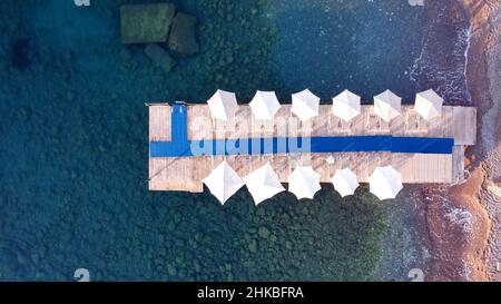 Vista dall'alto del molo di legno con ombrelloni e lettini da mare nella località turistica. Acqua di mare trasparente. Rocce sul fondo del mare. Mare calmo da drone vista Foto Stock