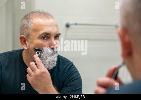 Uomo maturo dai capelli grigi che restyling la sua barba in casa utilizzando il rasoio Foto Stock